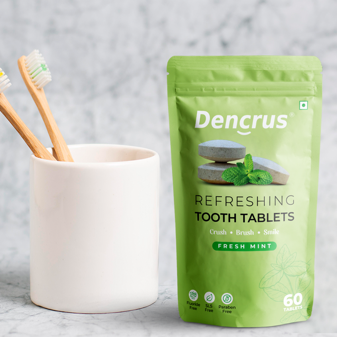 Dencrus Fresh Mint Tooth tablets are placed on white surface and beside there is a cup on which two toothbrushes are placed against a white textured background.