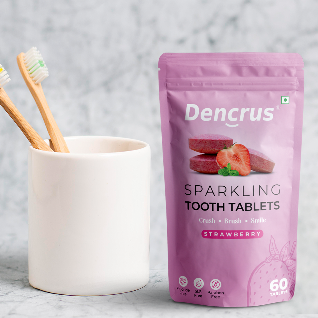 A package of Dencrus Sparkling Strawberry tooth tablets is placed against a textured and blurry white background. Alongside with the packaging there are two toothbrushes on a cup