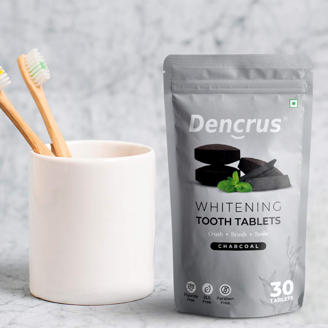 A package of Dencrus whitening tooth tablets with charcoal flavor displayed with two toothbrushes in a white mug against a white marble background