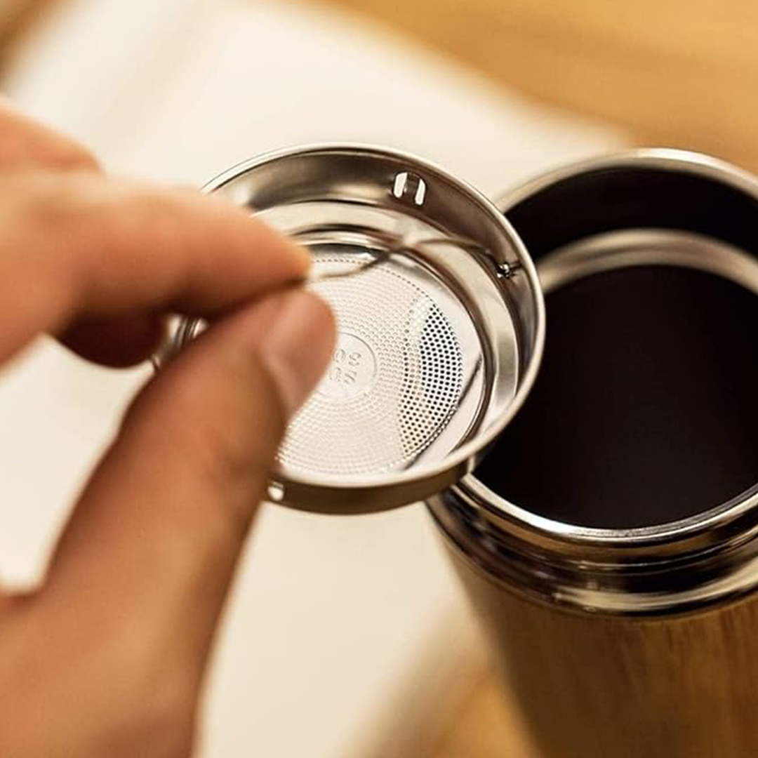 A human hand is holding the strainer of the IMECO Bamboo Flat lid Bottle while the rest of the bottle is visible and placed on a wooden surface