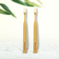 Two Dencrus NANO bamboo toothbrushes with white bristles standing upright on a wooden surface, with the brand name 'Dencrus' visible on the handle.