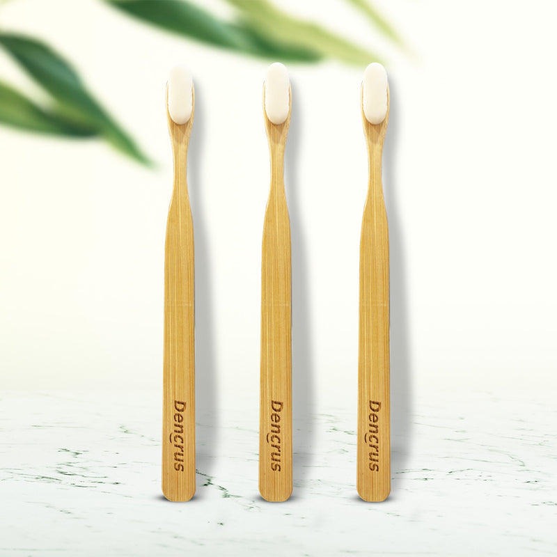 Three NANO bamboo toothbrushes with white bristles standing upright on a wooden surface, with the brand name 'Dencrus' visible on the handle.