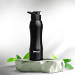 A black IMECO stainless steel water bottle with a flip-top lid, branded 'imeco', displayed on a green, leaf-strewn surface.