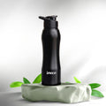 A black IMECO stainless steel water bottle with a flip-top lid, branded 'imeco', displayed on a green, leaf-strewn surface.