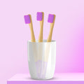 Three ORBY Kids bamboo toothbrushes with lavender bristles standing upright in a white cup, set against a pink background.